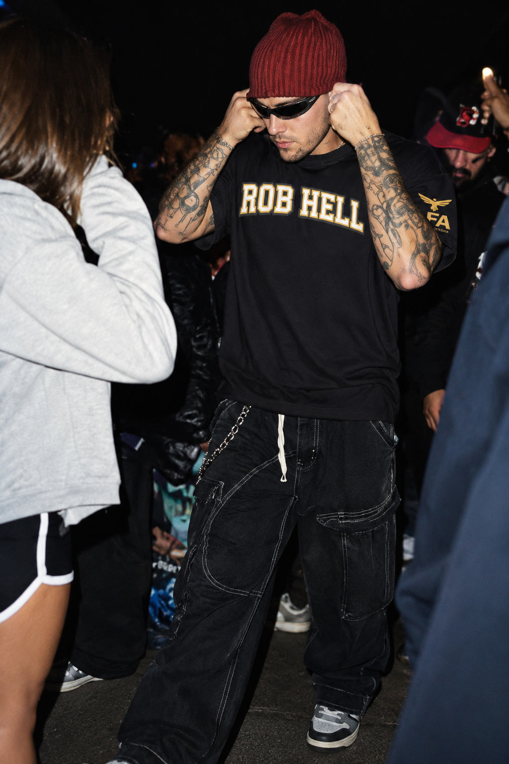A man in a crowd at night wearing a black t-shirt with 'ROB HELL' printed in gold on the chest, black cargo pants, a dark red knit beanie, and sunglasses.