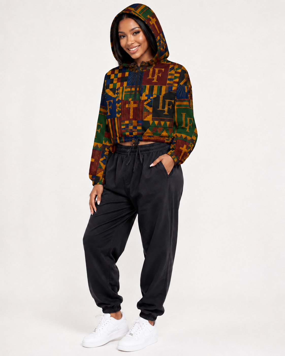 A woman modeling a cropped long-sleeve hoodie with a colorful geometric pattern in green, yellow, orange, and brown, paired with black jogger sweatpants and white sneakers, against a plain white studio background.