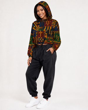 A woman modeling a cropped long-sleeve hoodie with a colorful geometric pattern in green, yellow, orange, and brown, paired with black jogger sweatpants and white sneakers, against a plain white studio background.