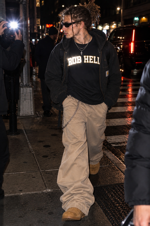 A man walking on a city sidewalk at night wearing a black hoodie with 'ROB HELL' printed in beige across the chest, beige cargo pants, and tan boots.