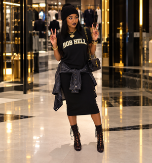 A woman posing in a luxury mall setting wearing a black crewneck t-shirt with 'ROB HELL' printed in gold, a black pencil skirt, a dark gray denim jacket tied around her waist, a black beanie, and black lace-up ankle boots.